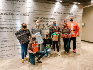 Seven individuals stand in front of a wall with DVIS logo and the names of DVIS donors. They are all masked, and each hold a present. Though you can't see their mouths, their eyes are definitely smiling!