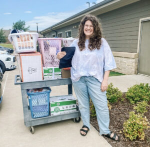 image description: a light-skinned woman with brown, curly hair stands in front of a rolling cart containing boxes and laundry baskets full of items. The woman is smiling and holding a clipboard.